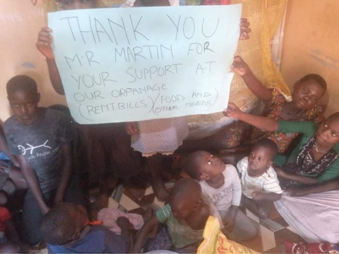 Children and caregivers holding a sign: thank you Mr Martin for your support at our orphanage — rent, bills, food and other needs